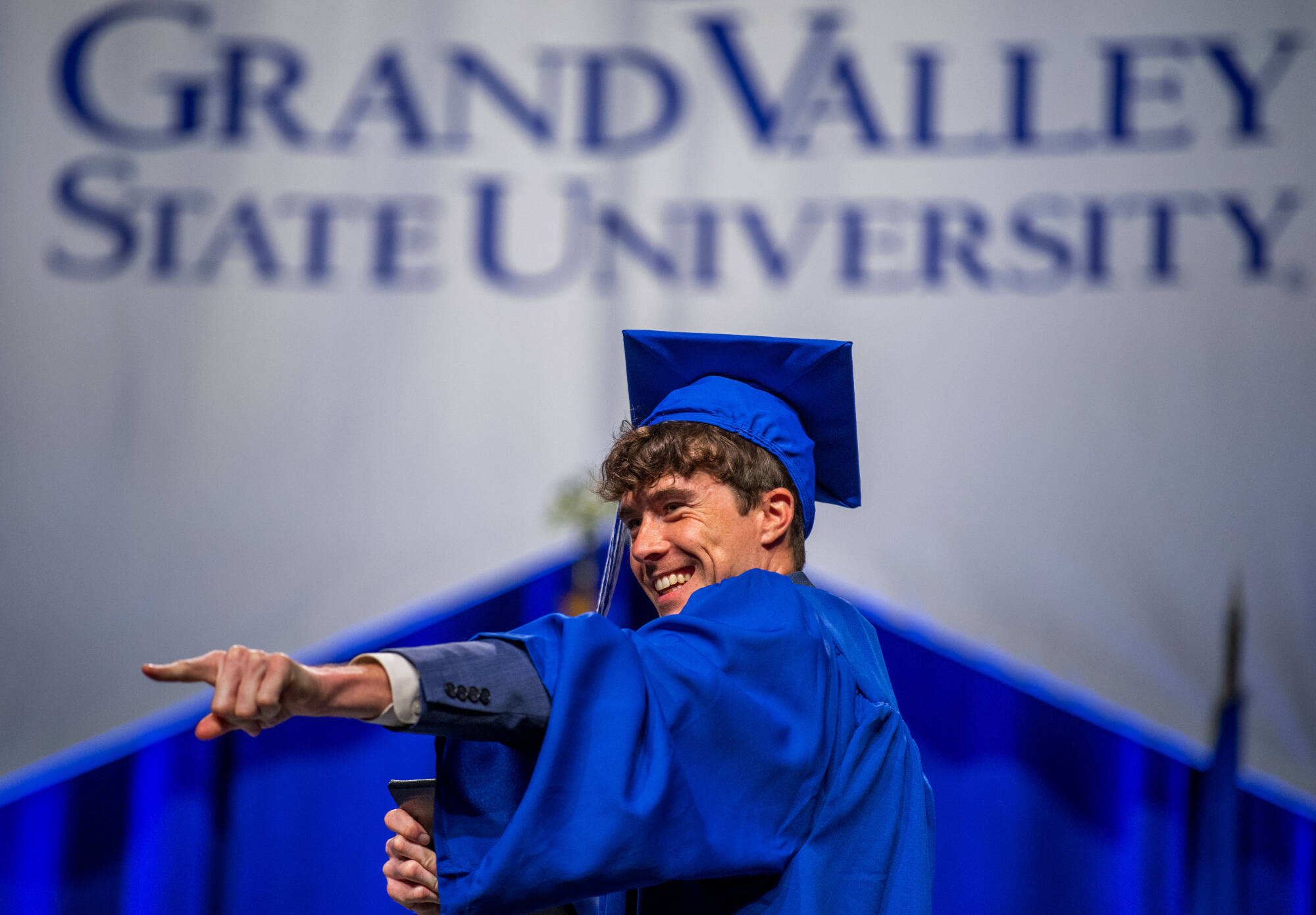 student wearing commencement robes, pointing out to a crowd, smiling, on stage to receive diploma